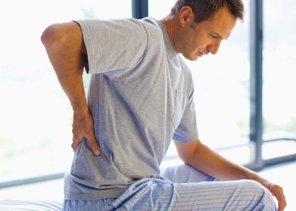 A man sits on the edge of his bed and rubs his lower back in pain.