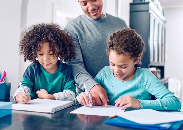 A father helps his two young children with their homework at the kitchen counter.