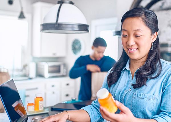 A woman checks the label on her prescription pill bottle in her kitchen while her husband unloads groceries in the background.