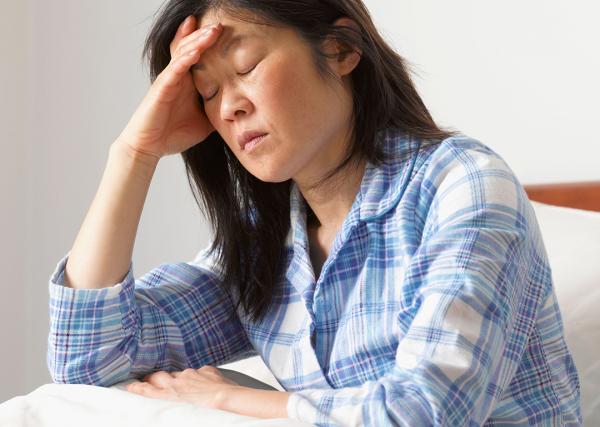 A woman suffering from a migraine sits up in bed while closing her eyes and holding her head.