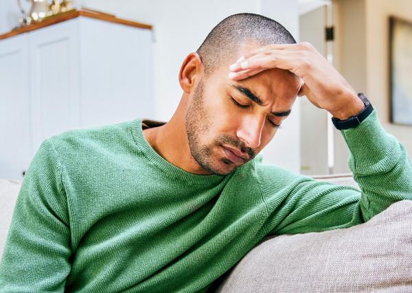 A man with a headache sits on his couch with his eyes closed, holding a hand to his head.