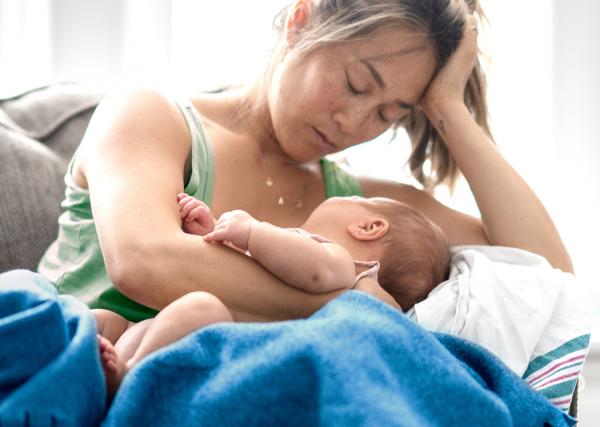 A mother sits on her couch feeling overwhelmed as she holds her newborn baby.