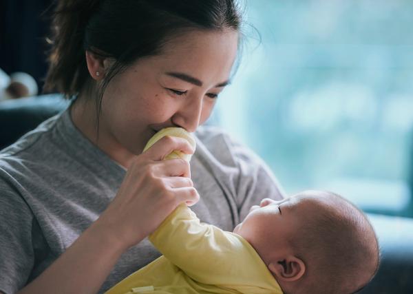 A smiling mother holds her young baby and kisses the baby’s hand.