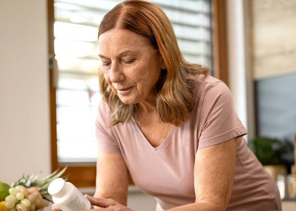 An older woman reads the label on an over-the-counter medication bottle in her kitchen.]