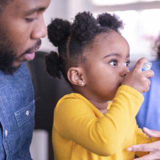 A young child sitting on her father’s lap uses an inhaler with the help of a healthcare provider.