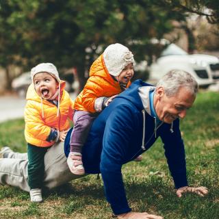 An older man does pushups outside with his two young grandchildren sitting on his back.