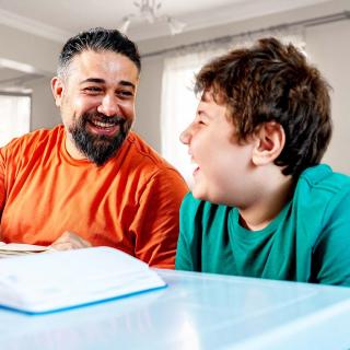 A father and his young son laugh and smile while the father helps his son with homework.